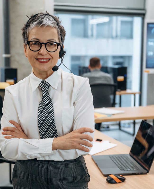 Businesswoman wearing a headset is smiling confidently in a modern office setting, emphasizing teamwork and professional communication with colleagues engaged in the background